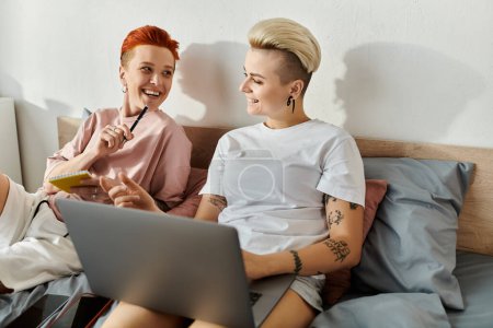 Two women with short hair sitting on a bed, engaged in using a laptop together.