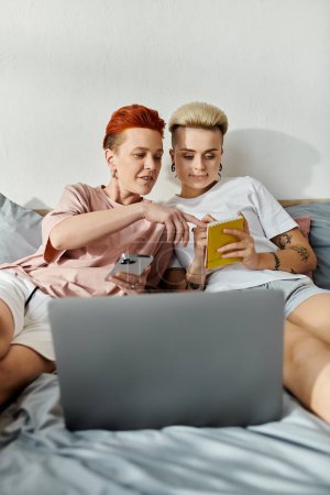 Two women, a lesbian couple with short hair, sit on a bed in a bedroom, focused on a laptop screen together.
