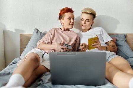 A lesbian couple with short hair sitting on a bed, focused on using a laptop in their bedroom.