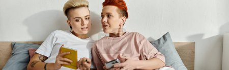 Téléchargez les photos : Un couple lesbien aux cheveux courts assis près d'un lit, tenant un téléphone portable, engagé dans un moment chaleureux et intime. - en image libre de droit