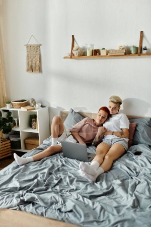 A lesbian couple with short hair sitting on a bed, immersed in their laptop screen, showcasing a modern LGBT lifestyle.