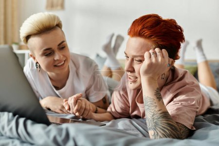 Foto de Dos mujeres con el pelo corto yacen en una cama, absortas en una pantalla de ordenador portátil, compartiendo un momento acogedor e íntimo. - Imagen libre de derechos
