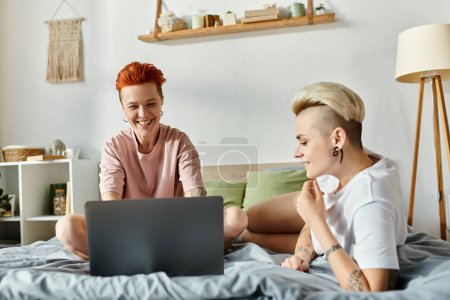 Foto de Una pareja lesbiana con el pelo corto disfrutando de la compañía de los demás en una cama mientras usa un ordenador portátil. - Imagen libre de derechos