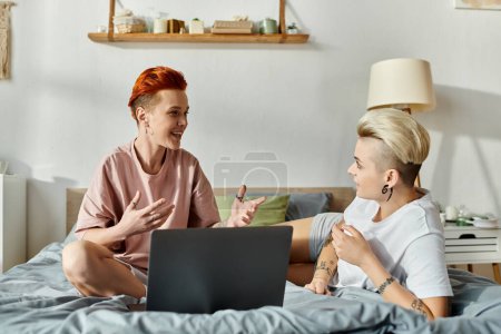 A lesbian couple with short hair sitting on a bed, deep in conversation while using a laptop.