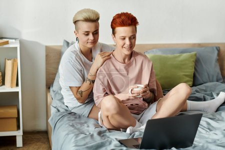 Foto de Dos mujeres con el pelo corto se sientan en una cama, absorto en una pantalla del ordenador portátil. - Imagen libre de derechos