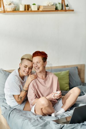 Zwei Frauen, ein lesbisches Paar, sitzen auf einem Bett mit Laptop und konzentrieren sich konzentriert auf ihre Arbeit im Komfort ihres Schlafzimmers.