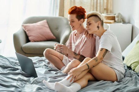 Lesbisches Paar mit kurzen Haaren sitzt eng auf dem Bett und ist in Laptop vertieft.