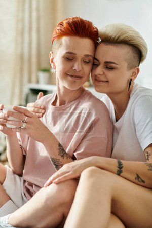 Two women with tattoos, one with a short haircut, sitting on a bed in a bedroom, showcasing their unique inked bodies.