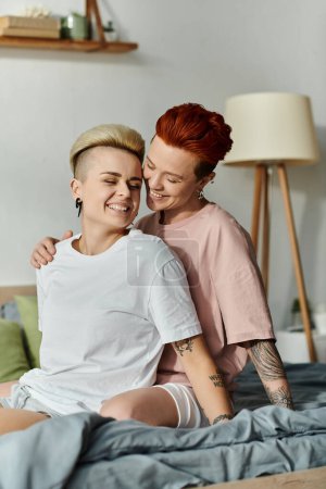 Téléchargez les photos : Un moment chaleureux capturé comme deux personnes, un couple de lesbiennes aux cheveux courts, assis sur un lit, s'embrassant dans leur chambre. - en image libre de droit