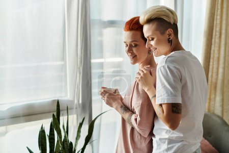 Photo for Two women, a lesbian couple with short hair, stand embracing in front of a window in their bedroom, showcasing their happy LGBT lifestyle. - Royalty Free Image