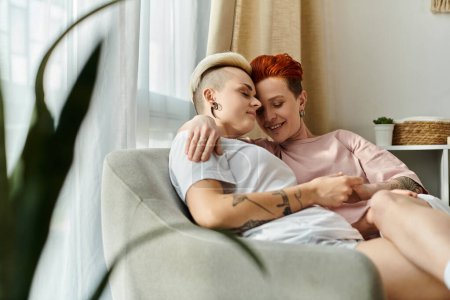 Foto de Un momento de corazón entre una pareja lesbiana, sentada en un sofá, abrazándose con amor en un acogedor dormitorio. - Imagen libre de derechos