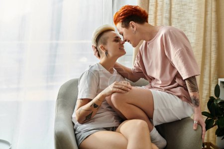 Foto de Dos personas, una pareja lesbiana de pelo corto, se sientan en una silla y comparten un beso en un momento íntimo. - Imagen libre de derechos