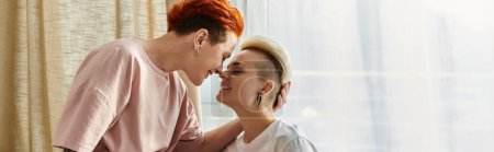 Foto de Dos mujeres con el pelo corto compartiendo un tierno beso delante de una ventana en un acogedor dormitorio, celebrando su amor en un momento íntimo. - Imagen libre de derechos