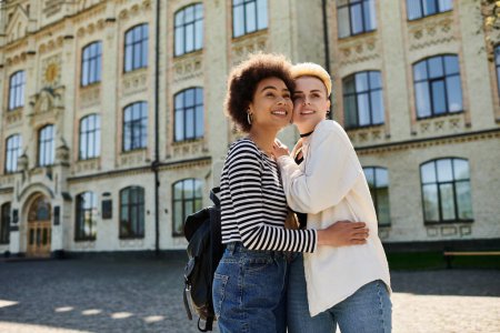 Two young women, one with light skin and the other with dark skin, embrace in a warm hug in front of a historic building.