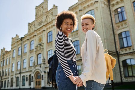 Dos mujeres jóvenes cogidas de la mano, de pie frente a un edificio moderno, compartiendo un momento de conexión y unión.