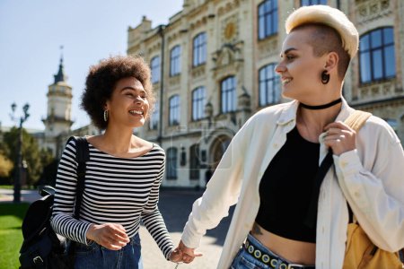 Dos mujeres jóvenes en atuendo de moda, caminan de la mano por una bulliciosa calle universitaria.