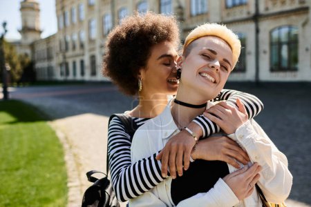 Foto de Una pareja lésbica multicultural, abrazada frente a un edificio universitario, mostrando amor y diversidad. - Imagen libre de derechos