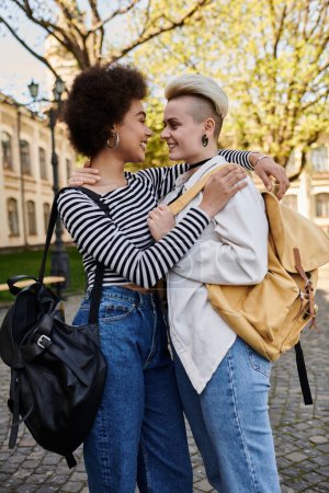 Téléchargez les photos : Deux jeunes femmes, une noire et une caucasienne, s'embrassent chaleureusement tout en tenant des sacs à dos à l'extérieur. - en image libre de droit