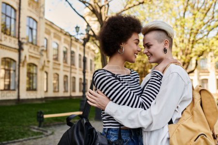 A multicultural lesbian couple hugs affectionately in front of a building on a university campus.
