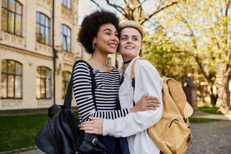 Dos mujeres jóvenes, una pareja lesbiana multicultural, abrazándose frente a un edificio en un campus universitario.