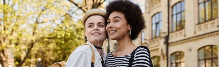 Un couple de lesbiennes multiculturel, vêtu élégamment, se tient ensemble dans une rue de la ville près d'un campus universitaire.