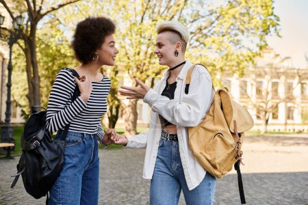 Two young women engaged in conversation in a vibrant park setting near a university campus.