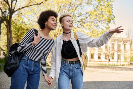 Foto de Dos mujeres jóvenes, una pareja lesbiana multicultural, se toman de la mano mientras caminan por una calle cerca de un campus universitario. - Imagen libre de derechos