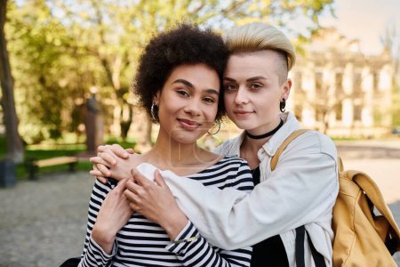 Two young women, one with dark hair and the other with blonde hair, share a warm hug in a lush green park.