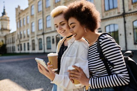 Téléchargez les photos : Deux jeunes femmes en tenue élégante, absorbées par un téléphone portable, se tiennent devant un bâtiment moderne dans une rue animée de la ville. - en image libre de droit