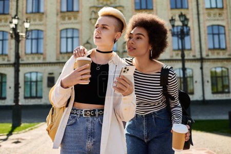 Two young women, a multicultural lesbian couple, stroll down a street near a university with coffee cups in hand.
