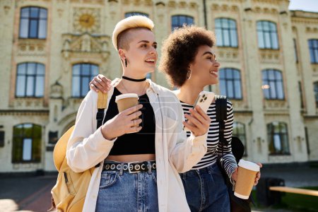 Foto de Dos mujeres jóvenes, una pareja lesbiana multicultural, pasean tranquilamente por la calle, cada una sosteniendo una taza de café. - Imagen libre de derechos