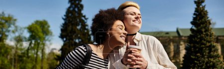 Two young women, a multicultural lesbian couple, stand in a park hugging each other warmly.