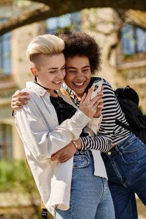 Two young women in casual attire embrace each other while engrossed in their cell phones, seemingly sharing a special moment.