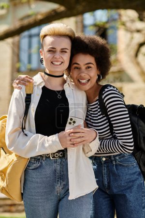 Dos mujeres jóvenes de pie frente a un campus, posando para una foto junto con sonrisas genuinas.