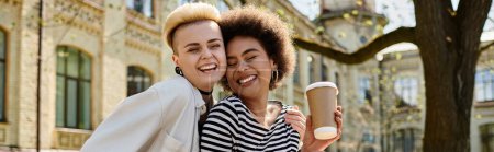 Junges, multikulturelles lesbisches Paar in stylischer Kleidung läuft die Straße entlang und hält Kaffeetassen in der Nähe des Universitätscampus.