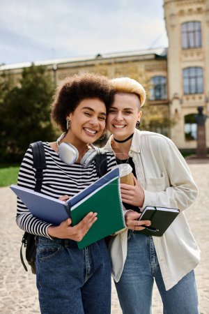 Two young women in casual attire, holding books, stand in front of a building on a university campus.