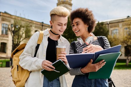 Téléchargez les photos : Un couple de lesbiennes multiculturel parle tout en tenant des carnets devant un immeuble sur un campus universitaire. - en image libre de droit