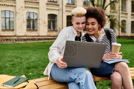 Two women in casual attire sitting on a bench, focused on a laptop screen, in an urban setting.