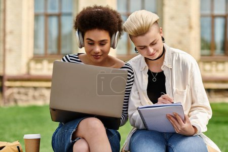 Dos mujeres jóvenes con atuendo casual sentadas en la hierba, centradas en el ordenador portátil.