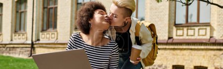 Téléchargez les photos : Deux jeunes femmes partagent un tendre baiser tout en tenant un ordinateur portable devant un bâtiment animé sur le campus. - en image libre de droit