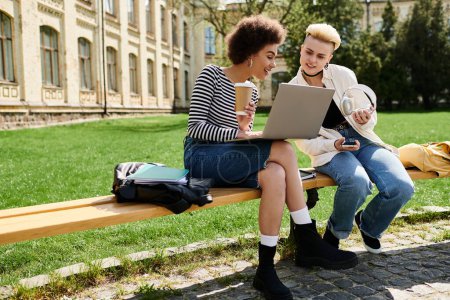 Téléchargez les photos : Deux femmes en tenue décontractée travaillant ensemble sur un ordinateur portable assis sur un banc à l'extérieur. - en image libre de droit