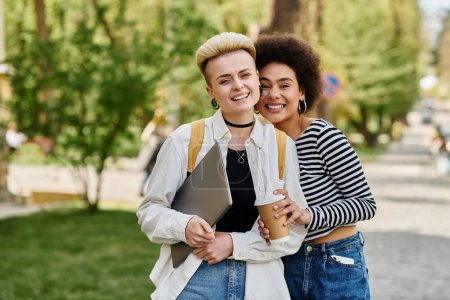 Foto de Dos jóvenes vestidas de manera casual se paran en un parque, sosteniendo tazas de café en un día soleado cerca de un campus universitario. - Imagen libre de derechos