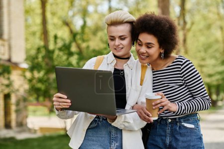 Foto de Dos mujeres jóvenes, casualmente vestidas, se sientan una al lado de la otra en un parque, enfocadas en una pantalla de computadora portátil, entablando una conversación. - Imagen libre de derechos