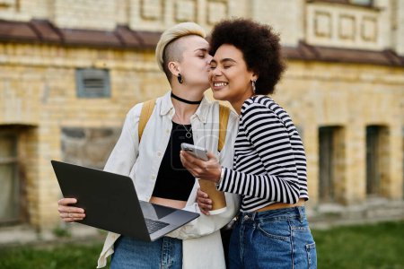 Téléchargez les photos : Deux jeunes femmes en tenue décontractée se tiennent devant un immeuble, travaillant sur un ordinateur portable. - en image libre de droit