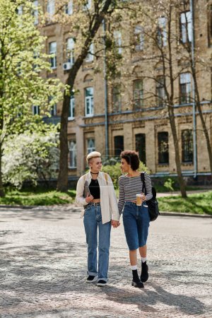 Foto de Dos mujeres jóvenes, una pareja lesbiana multicultural, paseando por una calle de la ciudad cerca de un campus universitario con un atuendo elegante. - Imagen libre de derechos