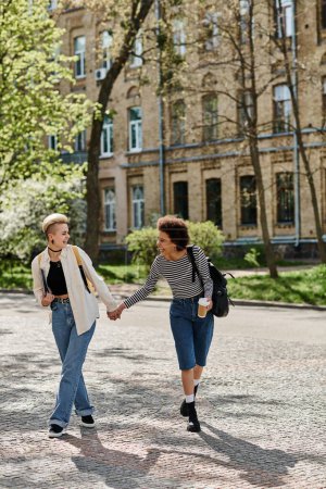 Foto de Dos mujeres jóvenes, cogidas de la mano, caminan por una calle empedrada cerca de un campus universitario. - Imagen libre de derechos