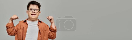 Foto de Un niño con síndrome de Down en gafas emana confianza con los brazos levantados en una pose poderosa. - Imagen libre de derechos