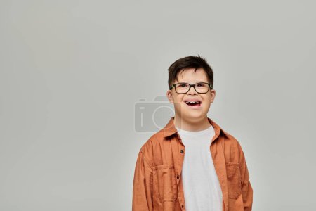 A little boy, with Down syndrome, wearing glasses, stands against a gray background.
