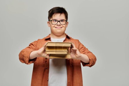 Un garçon trisomique tient joyeusement une pile de livres sur un fond gris.