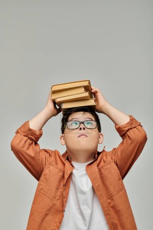 Foto de Un niño pequeño con síndrome de Down equilibra una pila de libros en su cabeza. - Imagen libre de derechos